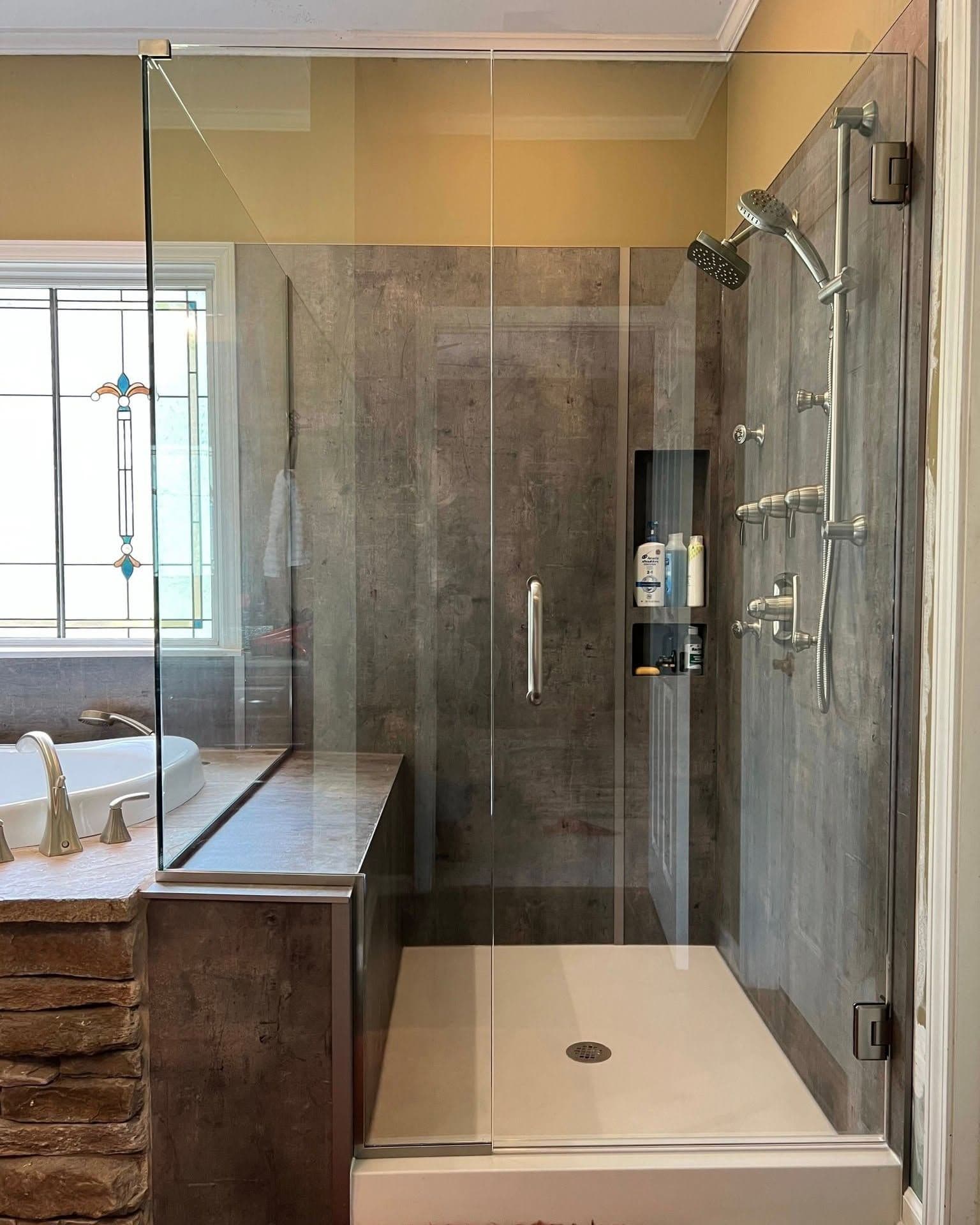 Luxury walk-in shower with gray tile walls, glass enclosure, rainfall showerhead, hand-held sprayer, and built-in niche with toiletries, adjacent to soaking tub and vanity in spa-like bathroom