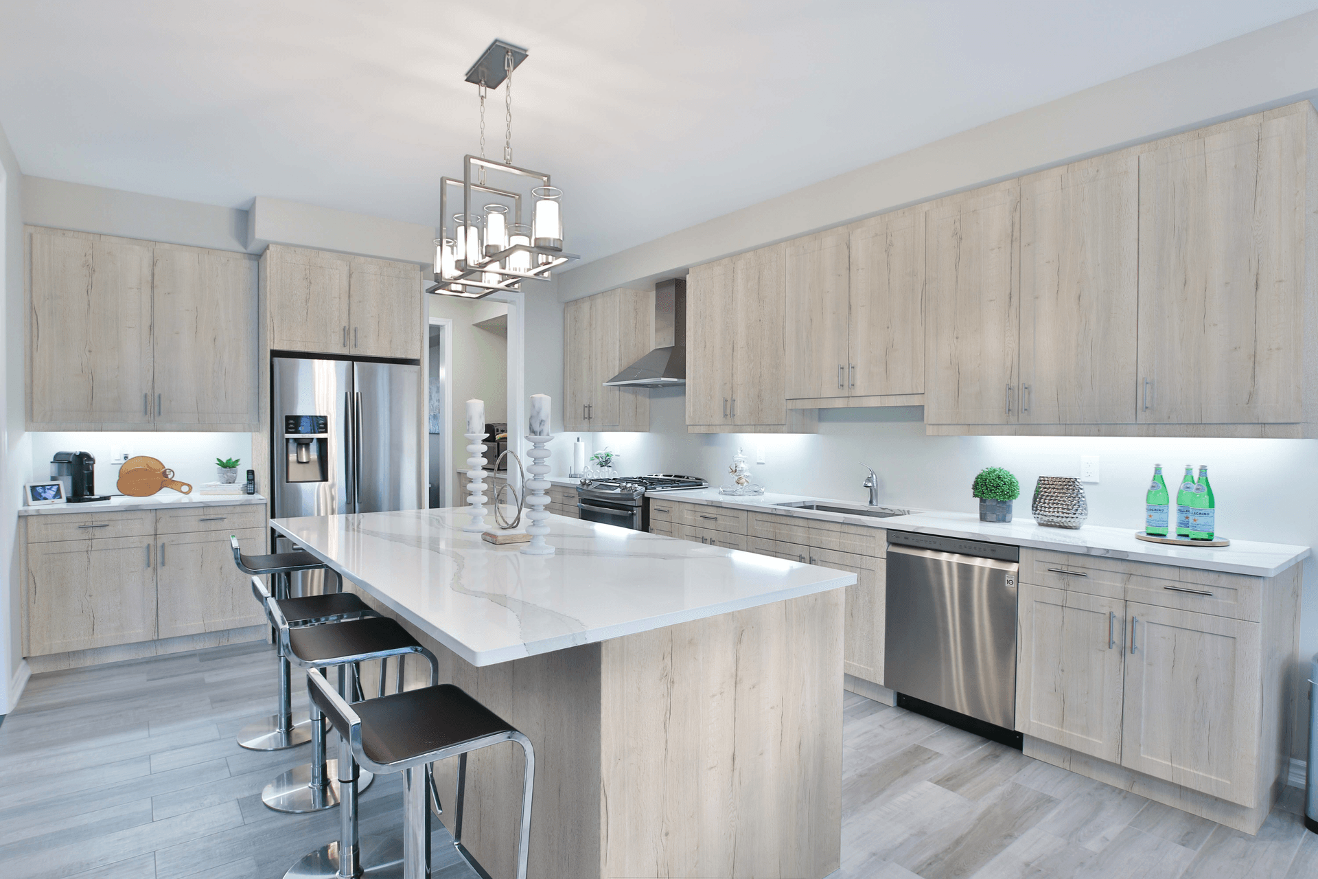 Modern kitchen with light wood cabinets, a large white island, stainless steel appliances, and bar stools. Bright lighting creates a clean, inviting atmosphere.