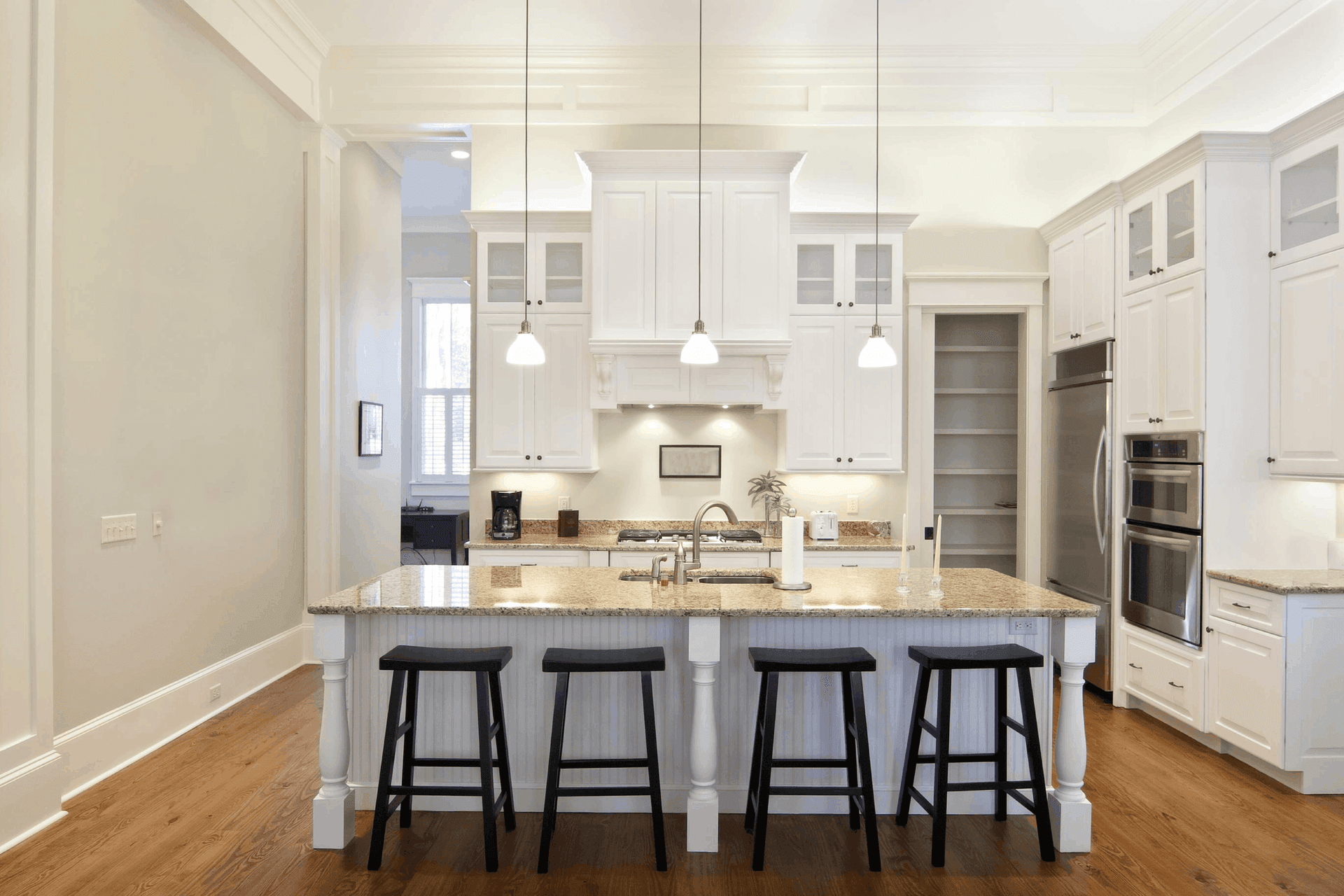 Spacious kitchen with white cabinets, granite countertops, and a large island with four black stools. Pendant lights hang above, creating a bright, inviting ambiance.