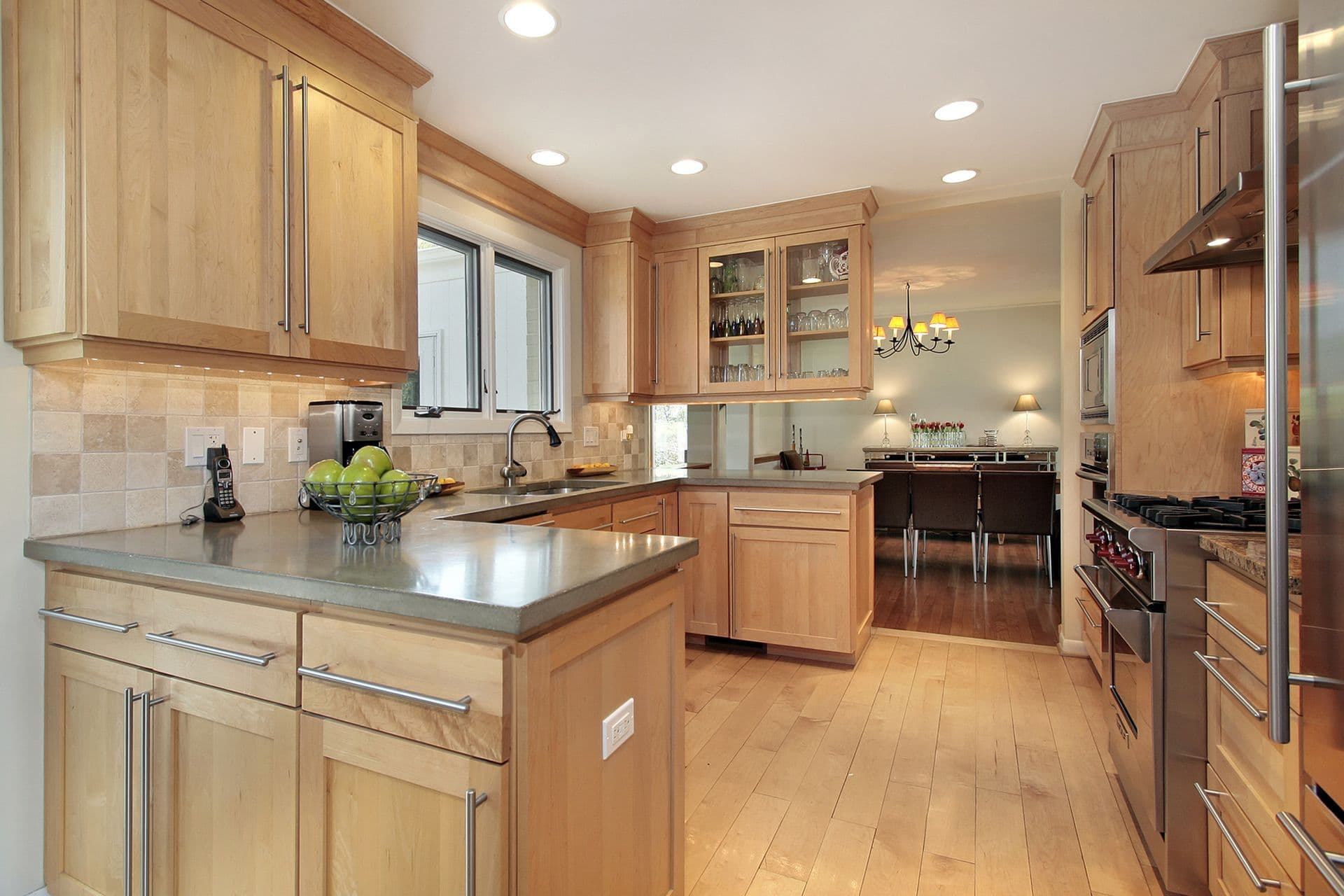 Modern kitchen with light wood cabinetry, gray countertops, and stainless steel appliances. Bright lighting and bowls of green apples create a fresh, inviting ambiance.