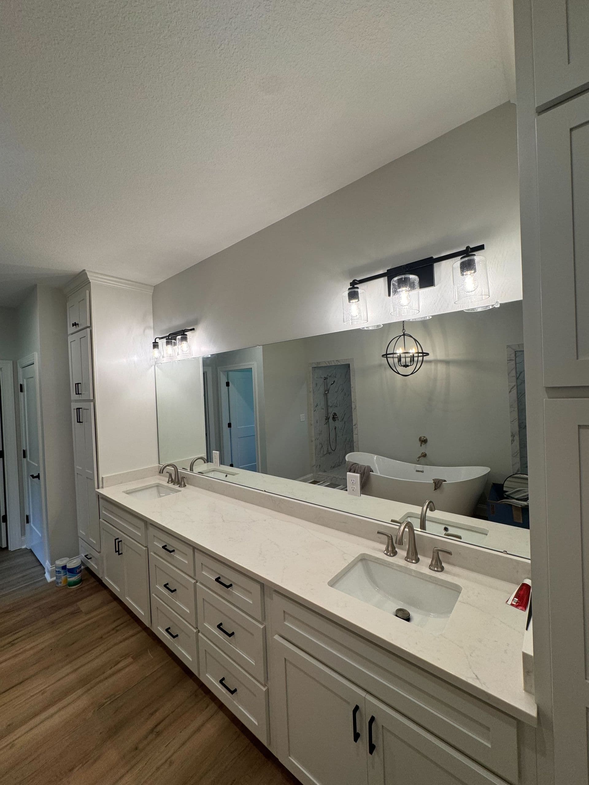 Spacious bathroom with a modern double sink vanity, large mirror, and industrial-style lights. Freestanding tub and wood floor add warmth.