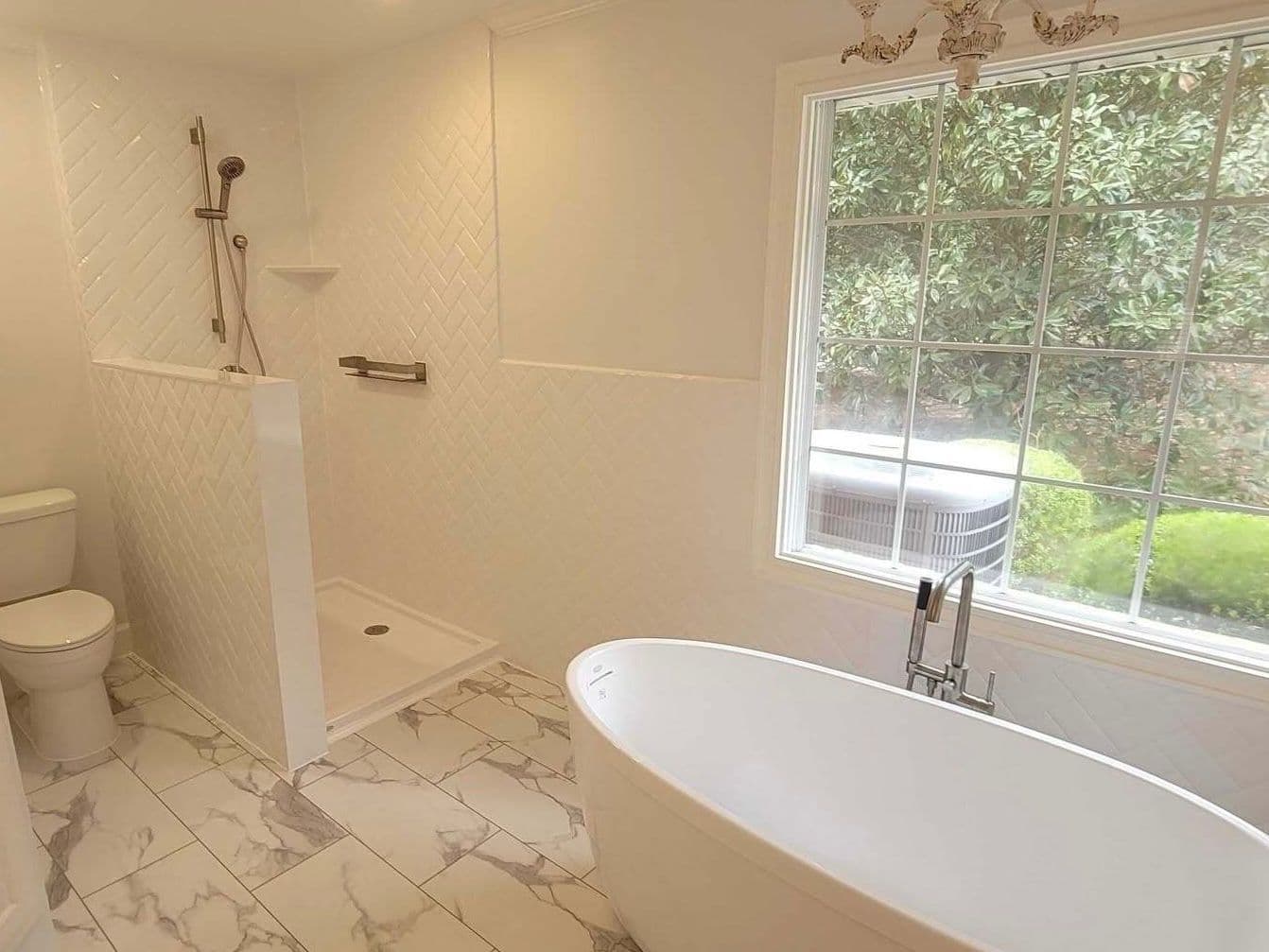 Modern bathroom with freestanding white tub, walk-in shower with herringbone tile, toilet, and large window overlooking green lawn and landscaping