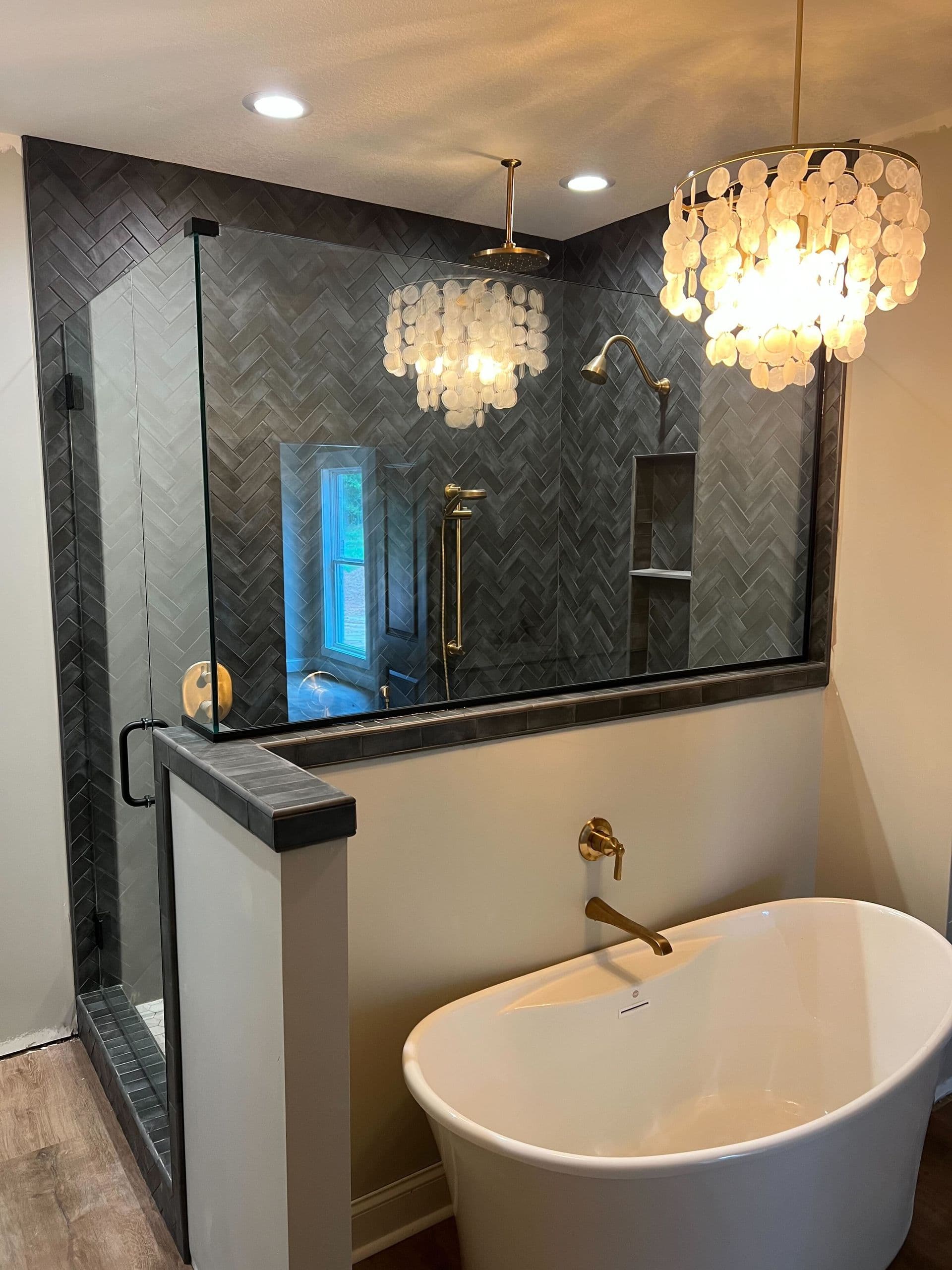 Modern luxury bathroom with freestanding white bathtub, walk-in shower with herringbone tile walls and glass enclosure, dual pendant chandeliers, and brass fixtures