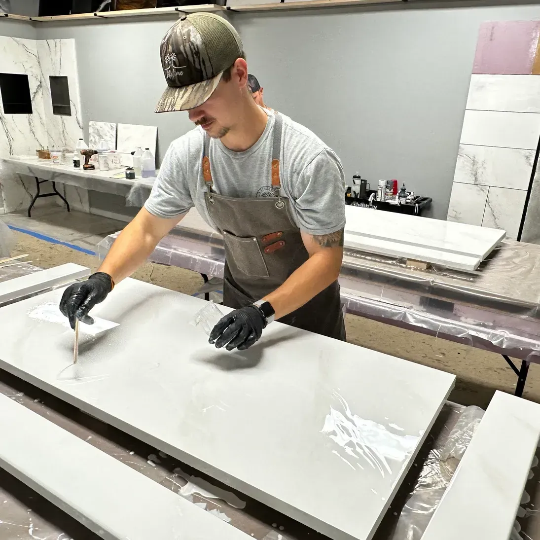 A person in a cap and apron uses a tool to work on a large, glossy countertop in a workshop. The setting conveys focus and craftsmanship.