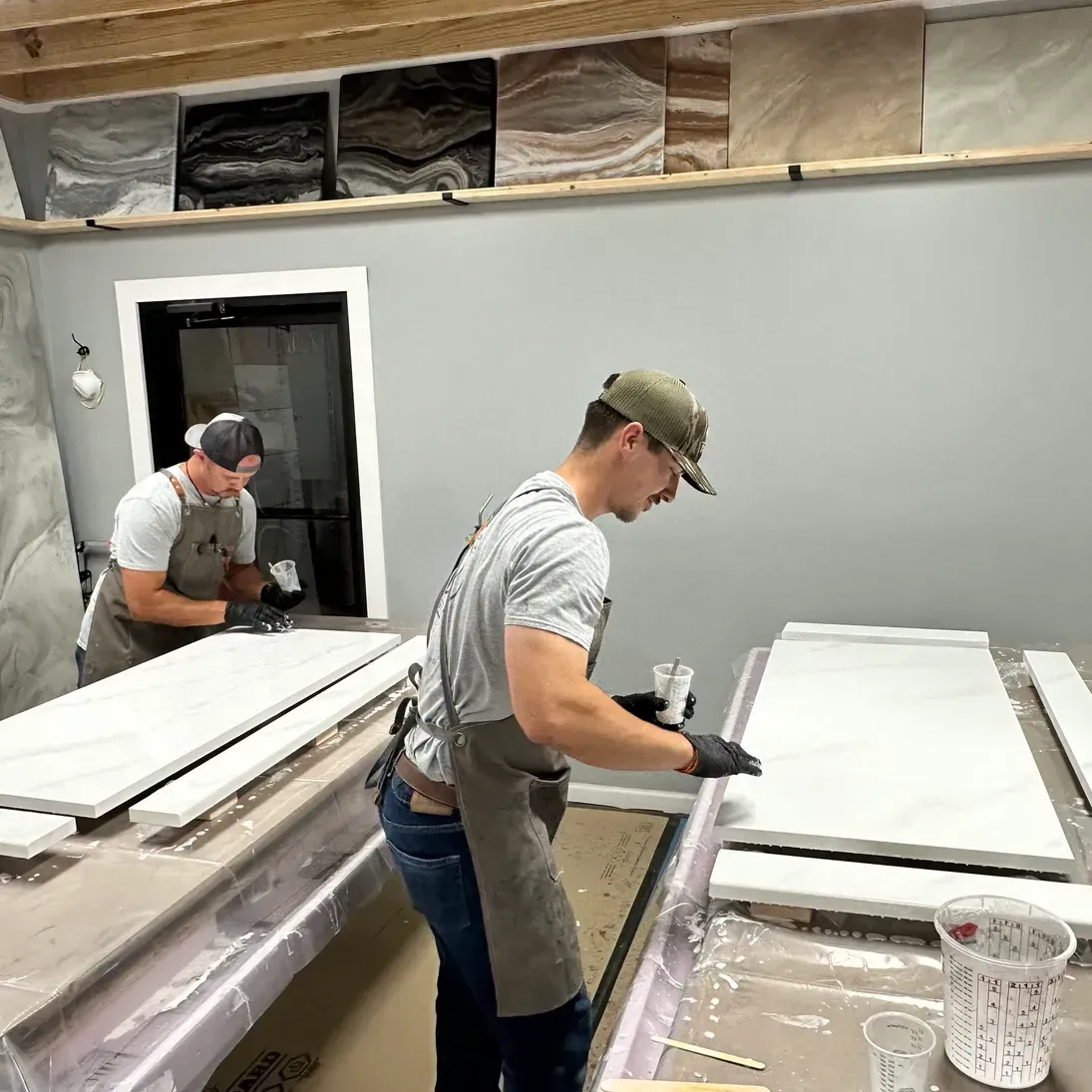 Two men in aprons and caps work on white stone slabs in a workshop. Sample stone panels are displayed on a wall. The setting is industrious.