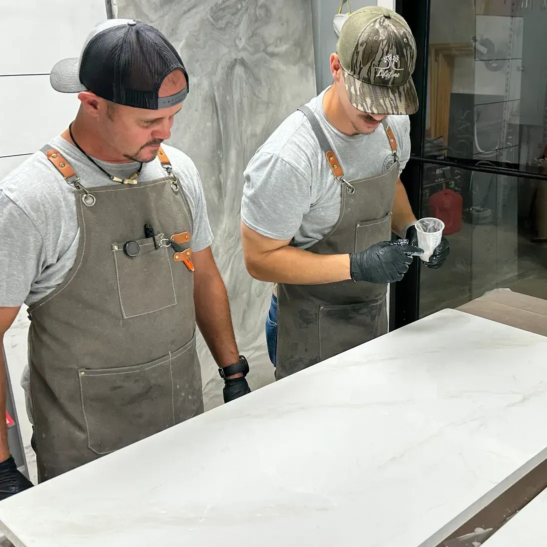 Two men wearing gray aprons and caps working on a large, smooth white countertop. One holds a small cup, focused and intent on their task.
