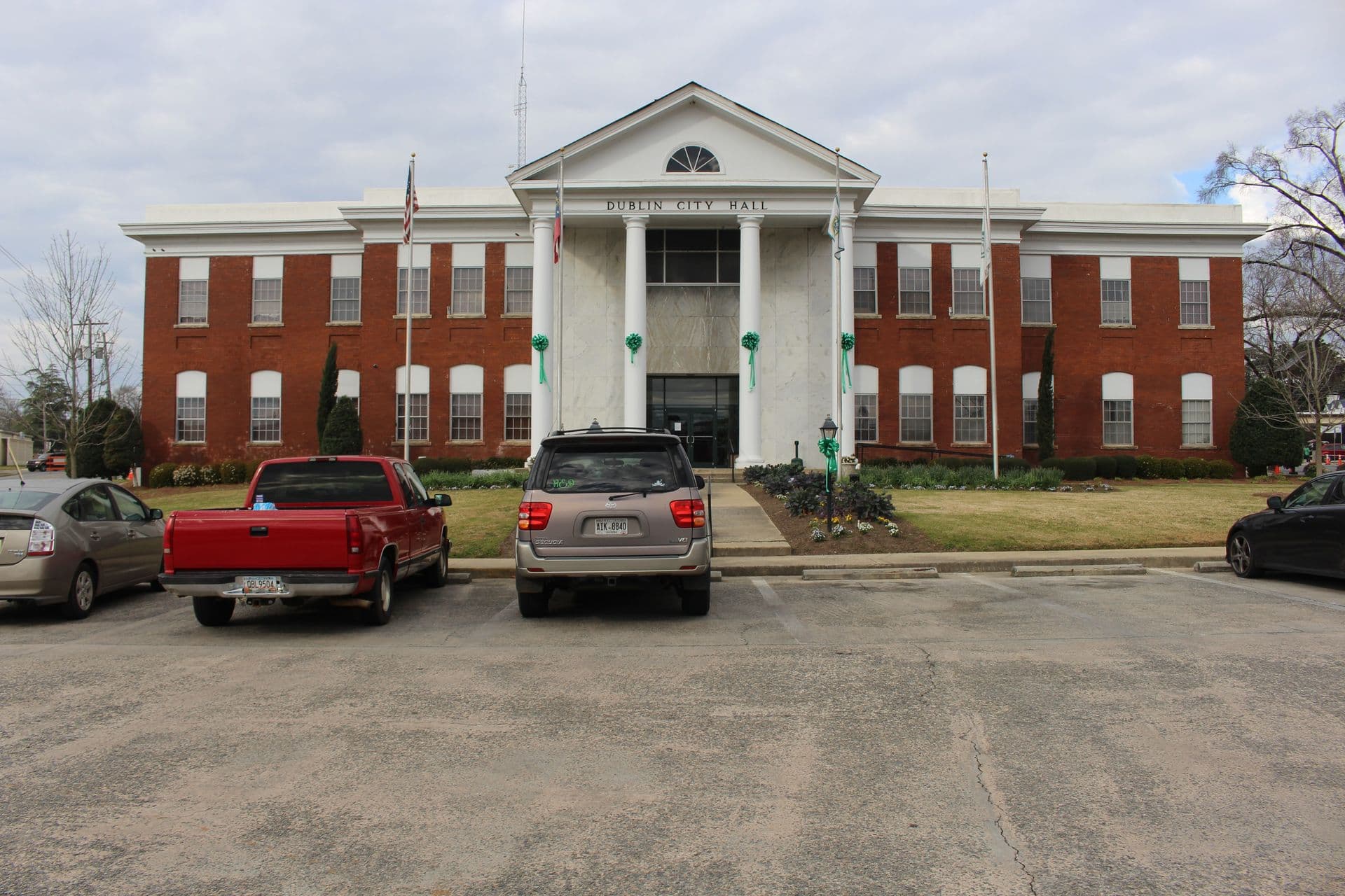 Red-brick city hall with white columns and "Dublin City Hall" sign, green decorations, U.S. flags. Three cars parked in front, cloudy sky.
