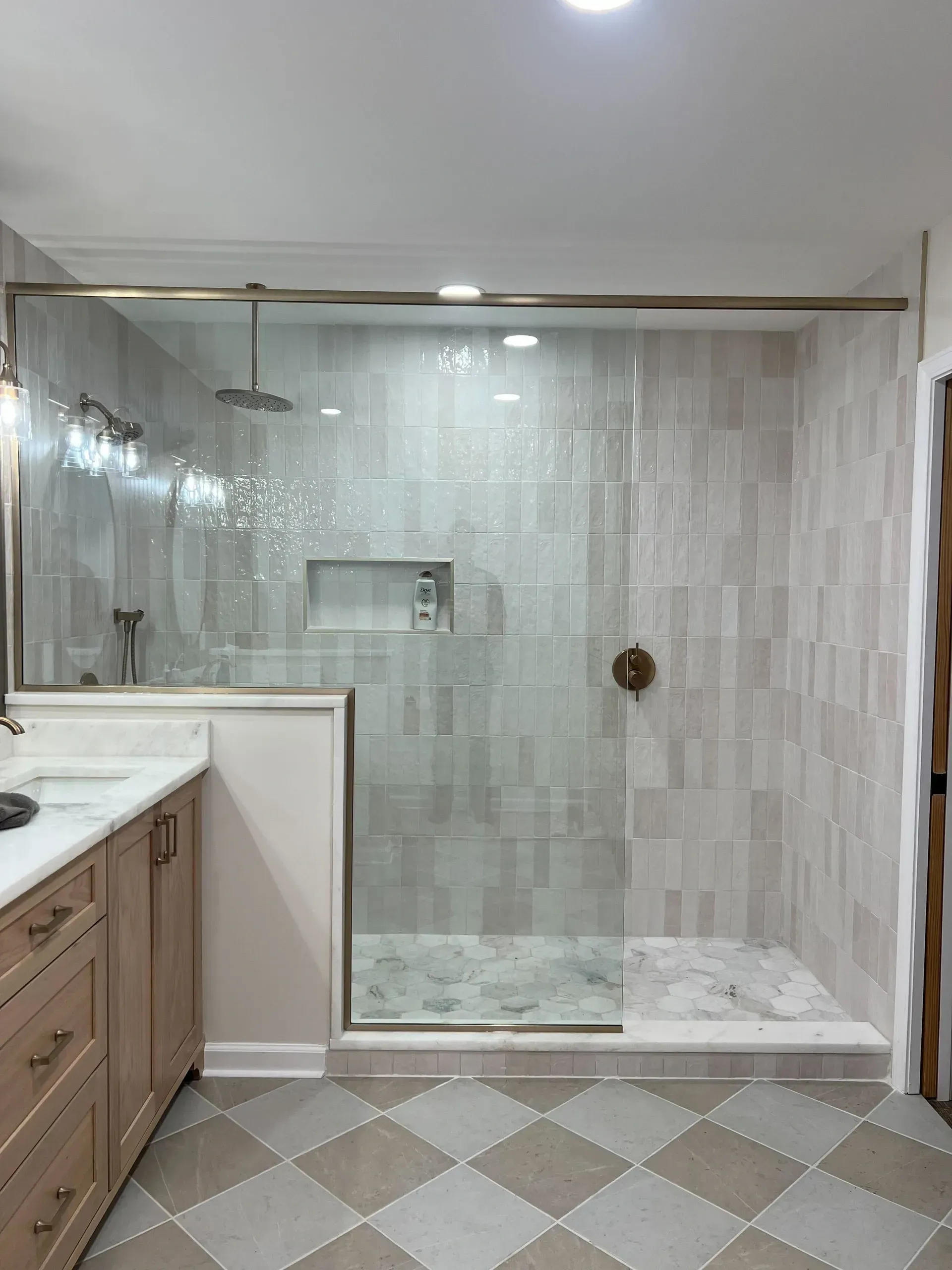 Modern bathroom with a large glass-enclosed shower. Features light beige tiles, a rainfall showerhead, marble countertop, and wooden cabinets.