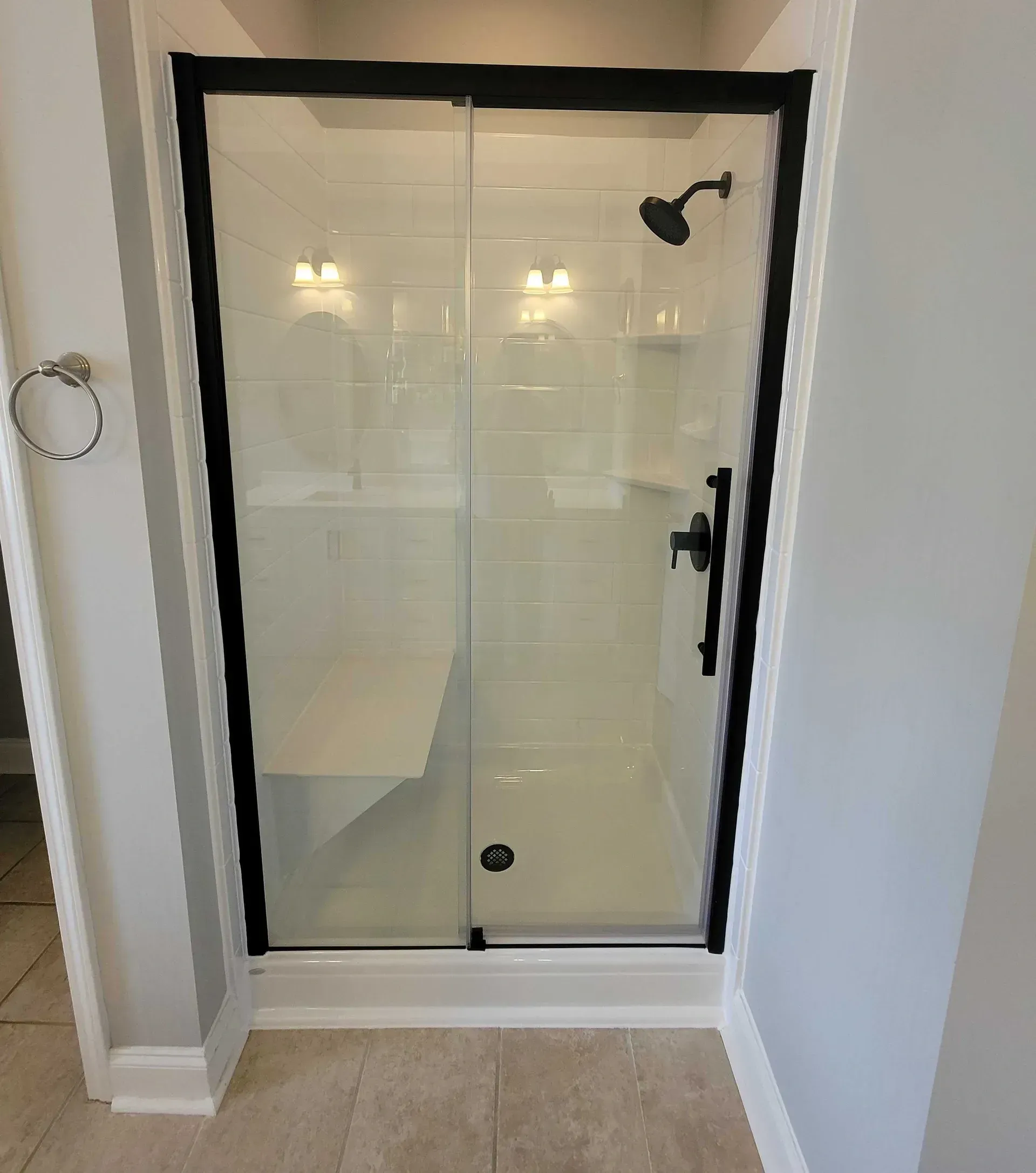 A modern shower with clear glass doors, black frame, and white tiled walls. It features a built-in bench, sleek black showerhead, and soft lighting.
