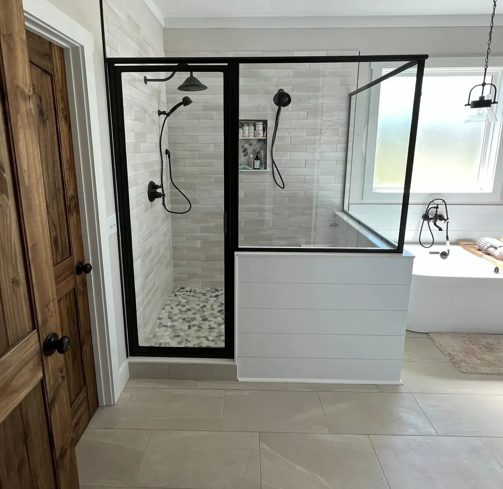 Modern bathroom with a large glass shower on the left, featuring black trim and beige tiles. On the right is a freestanding tub under a frosted window.