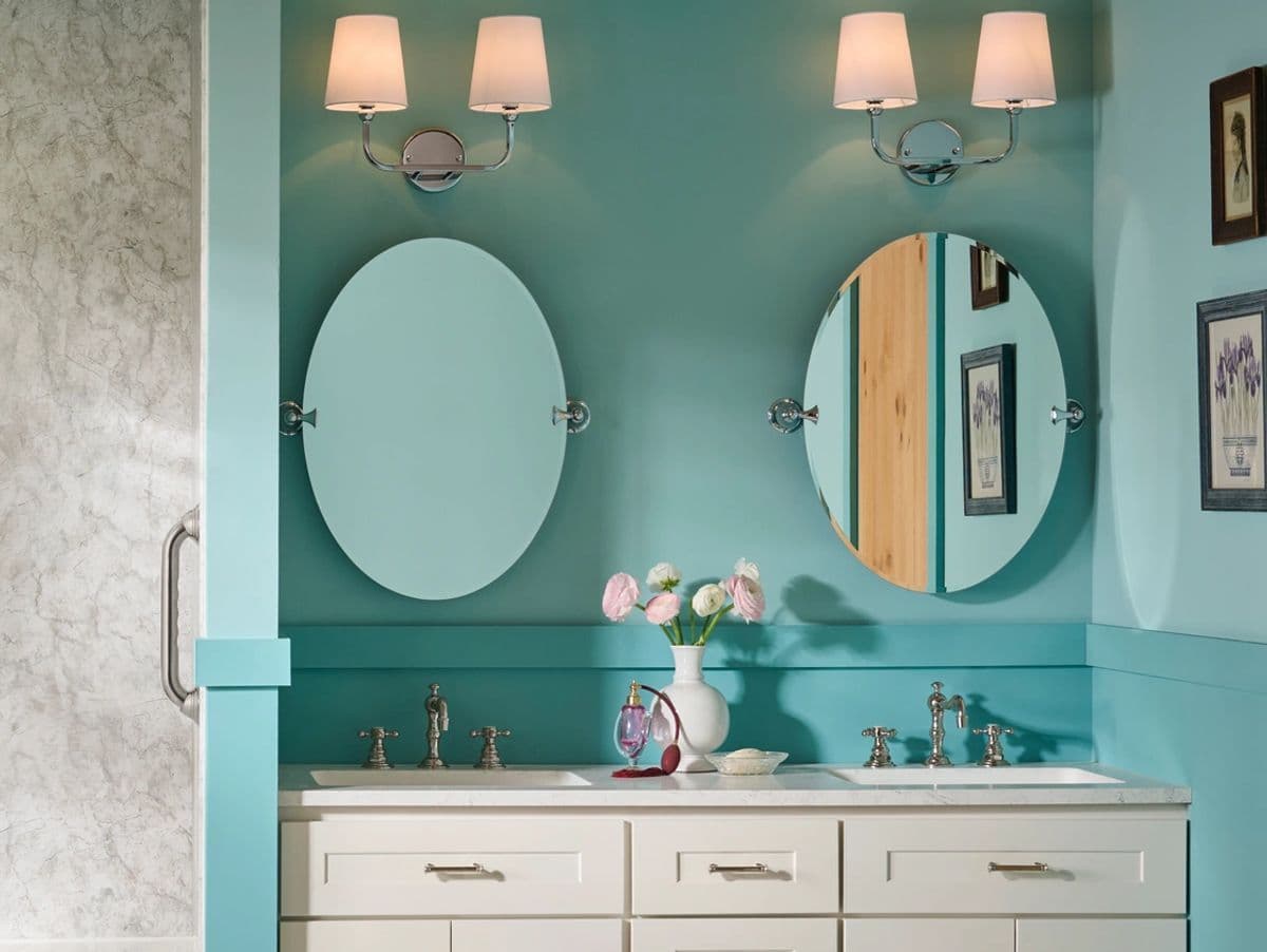 Elegant bathroom with light turquoise walls and two oval mirrors above a white double vanity. Soft lighting from two sconces and pink flowers in a vase add warmth.