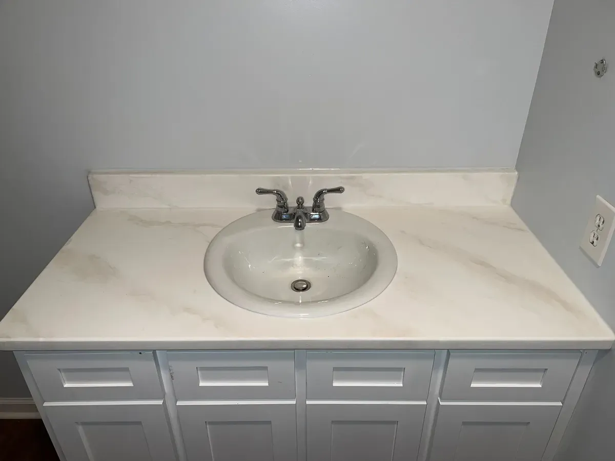 White bathroom vanity with a glossy marble countertop, round sink, and dual-handled faucet. Soft gray wall with an electrical outlet on the right.